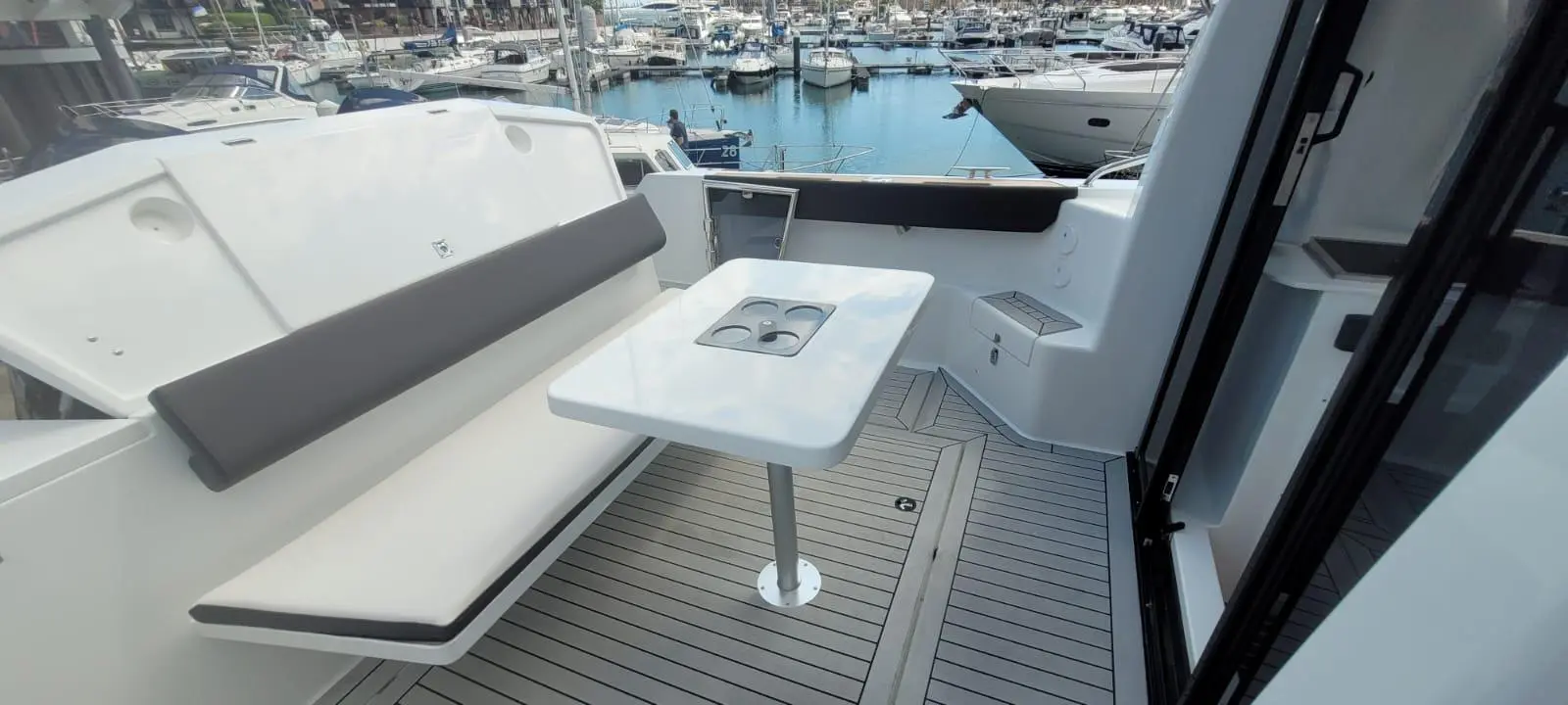 Interior of a yacht featuring a white table with cup holders, a gray cushioned bench, surrounded by boats in a marina.